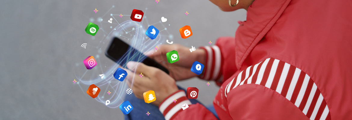 A imagem mostra uma pessoa com um casaco vermelho a utilizar um smartphone, do qual emergem vários ícones 3D de redes sociais (como WhatsApp, Instagram e YouTube) interligados por linhas de luz. A composição utiliza um fundo neutro para destacar a ideia de conectividade constante e a imersão no ecossistema digital.