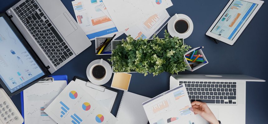 Ambiente de trabalho moderno com laptops, tablet e documentos com gráficos e análises financeiras espalhados sobre a mesa, acompanhado por café e elementos de escritório